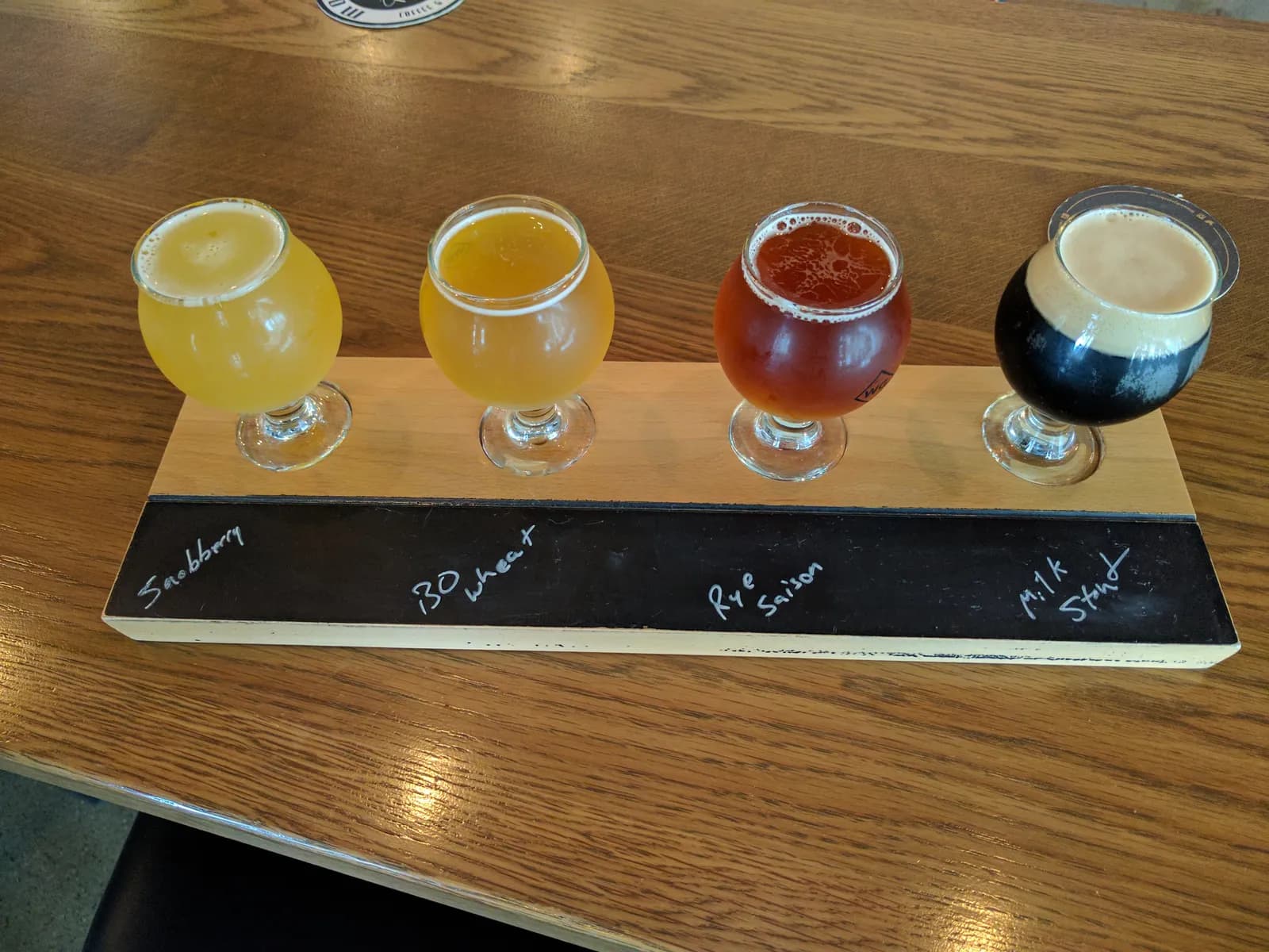 Planche de dégustation avec quatre verres à pied : deux bières claires, une ambrée et un stout crémeux, les styles écrits à la craie sur l'ardoise