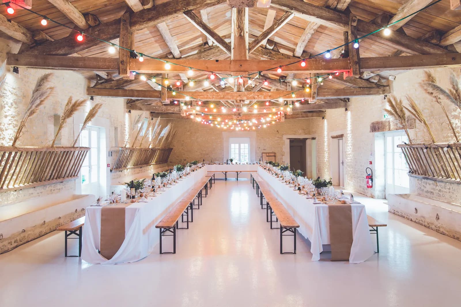 Salle de réception de mariage sous une charpente en bois, avec de longues tables nappées de blanc, des bancs et des guirlandes lumineuses