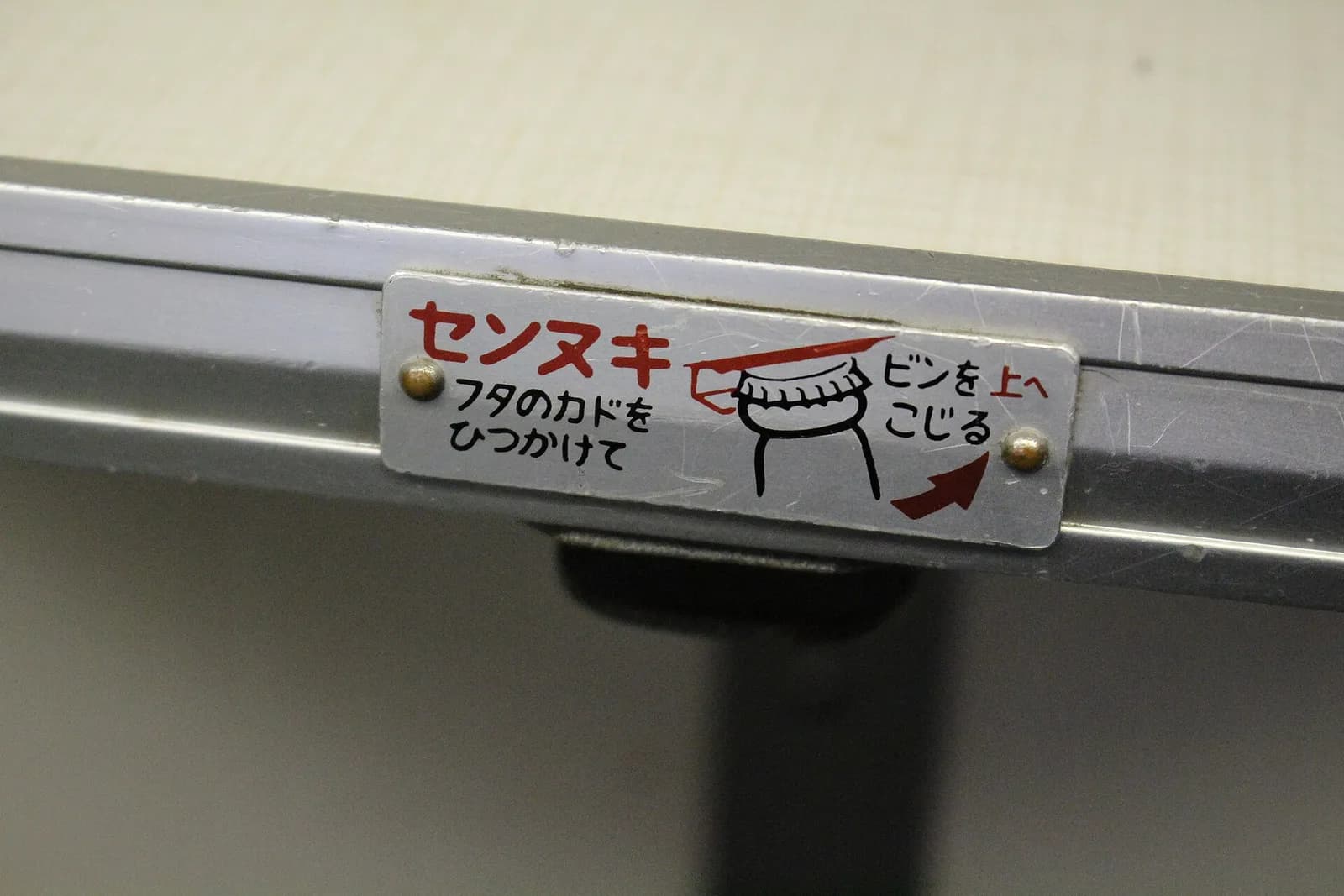 Décapsuleur fixé sous le rebord d'une tablette de train, avec une plaque métallique illustrée qui montre le geste pour ouvrir la bouteille