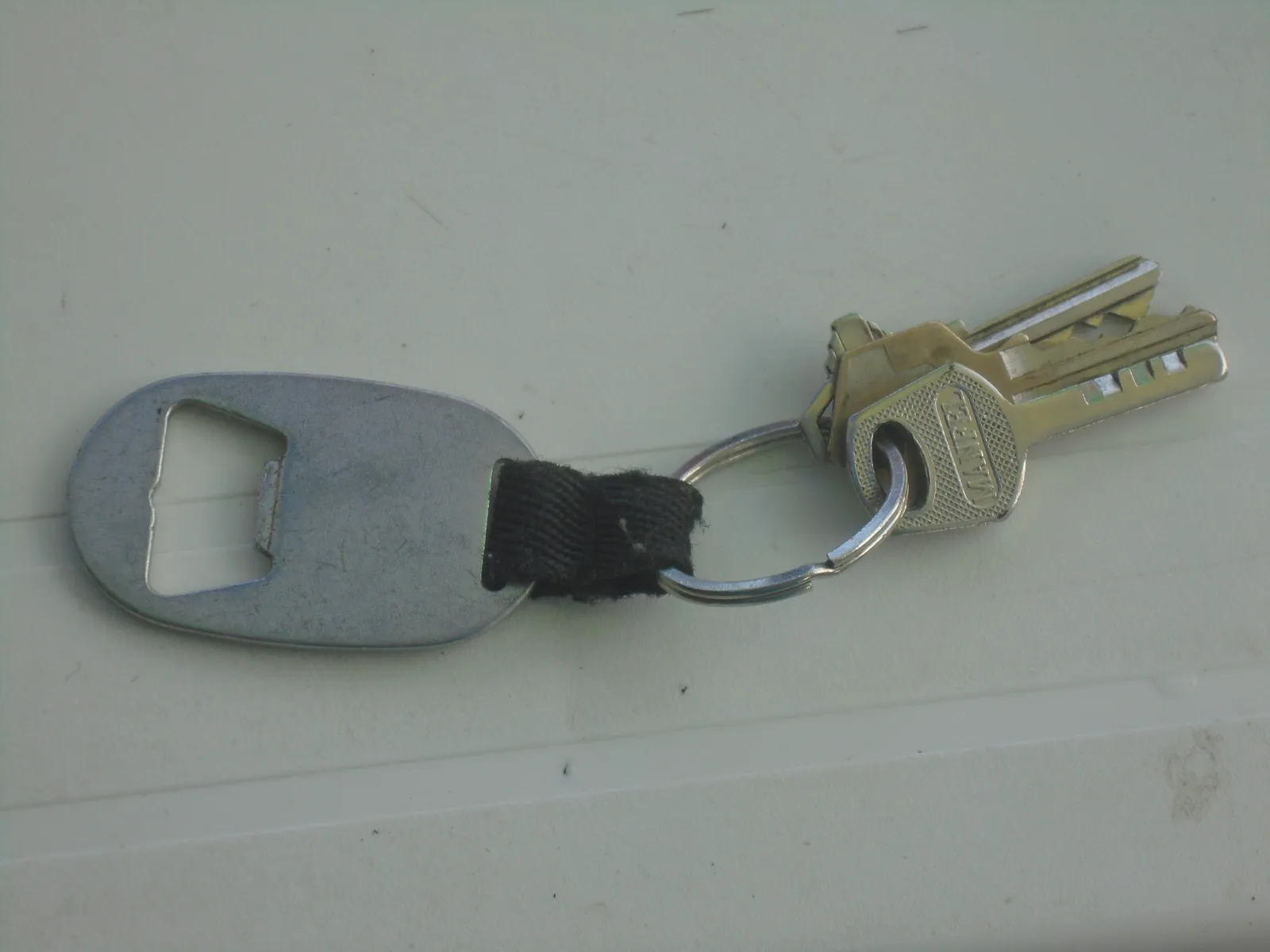 Décapsuleur plat en métal relié par une sangle noire à un anneau de porte-clés et à trois clés, posé sur une surface claire