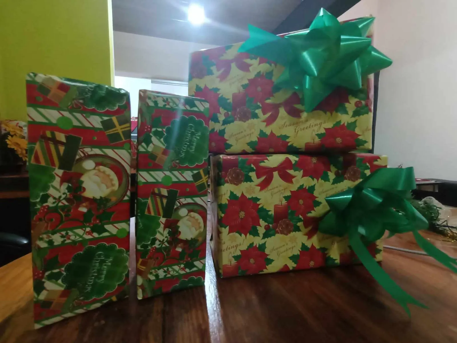 Quatre cadeaux de Noël emballés dans du papier à motifs de poinsettias et de Père Noël, avec des nœuds verts, posés sur une table en bois